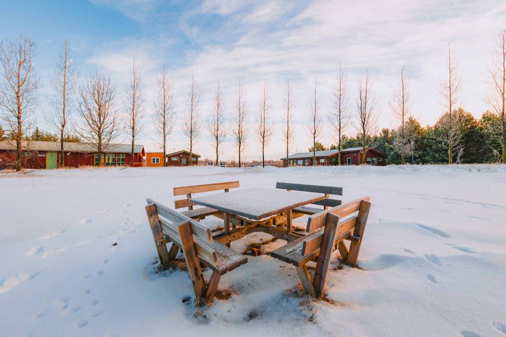 A table with four benches located in the middle of a snow-covered field next to the campground buildings.