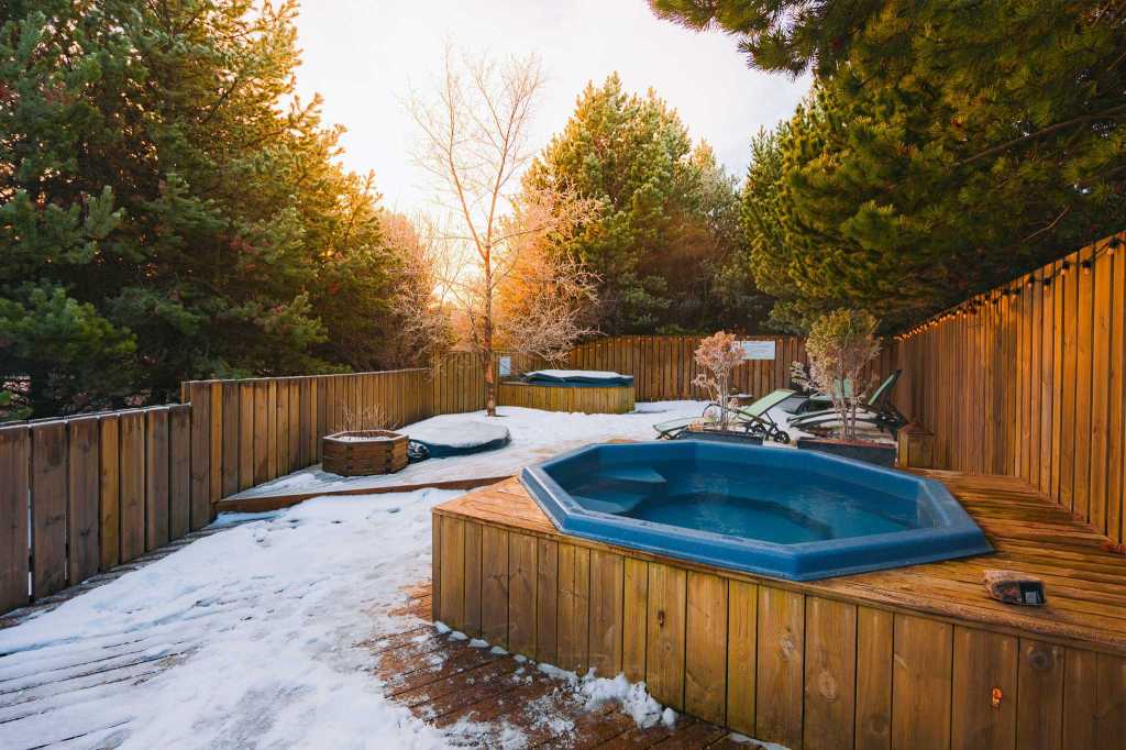A terrace surrounded by trees and a wooden fence, built of planks, with two hot pots and several deck chairs.