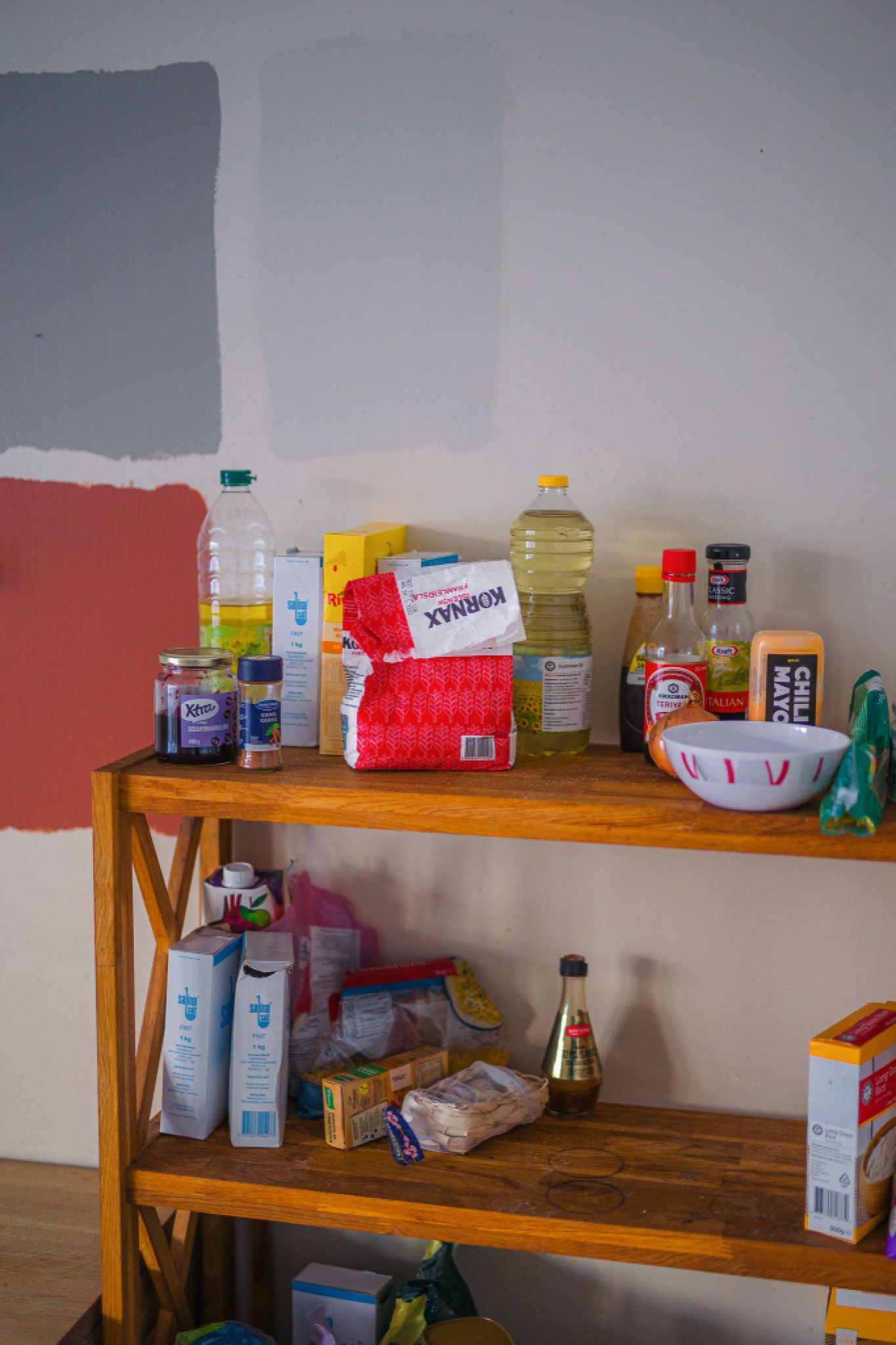 Wooden shelf against a wall with paint samples, holding various food items like oil, sauces, and cereal. The scene is casual and cluttered.