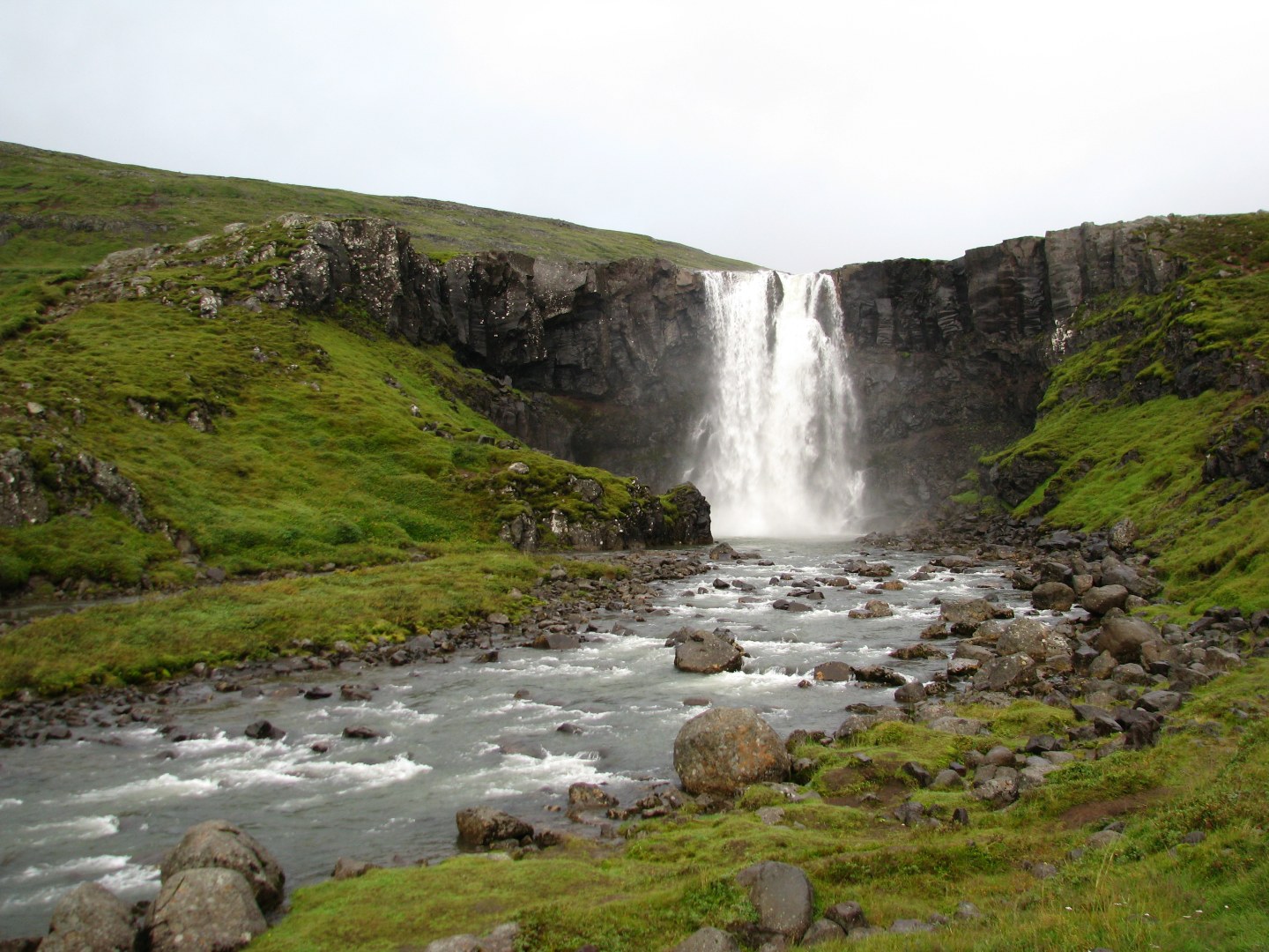 A stunning waterfall cascades over rugged cliffs, surrounded by lush green hills. A rocky river flows towards the foreground under a cloudy sky, evoking tranquility.