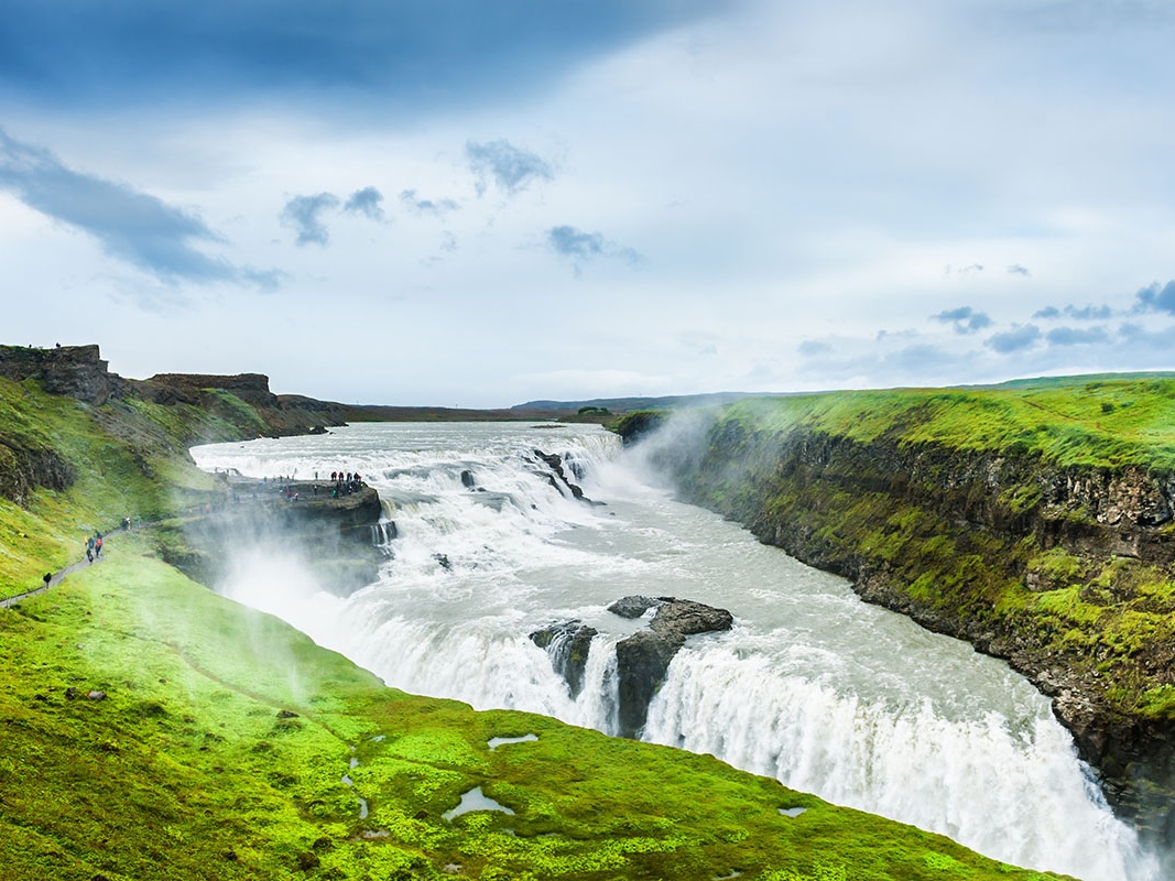 Majestic waterfall cascading over rocky cliffs, surrounded by vibrant green moss, under a cloudy blue sky. Tourists stand on a nearby viewing platform.