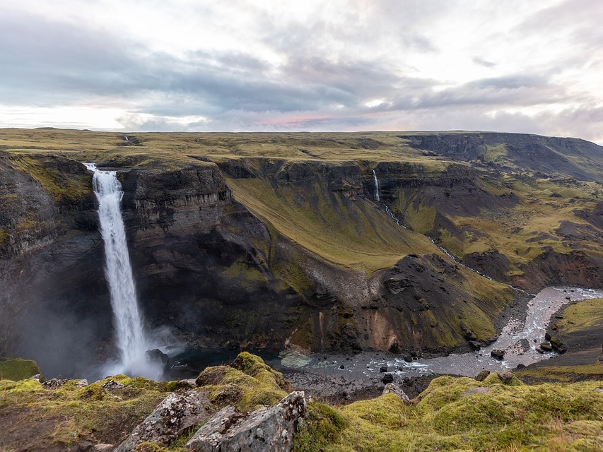 Majestic waterfall cascading off a rugged cliff into a river, surrounded by lush green hills under a cloudy sky, conveying tranquility and awe.