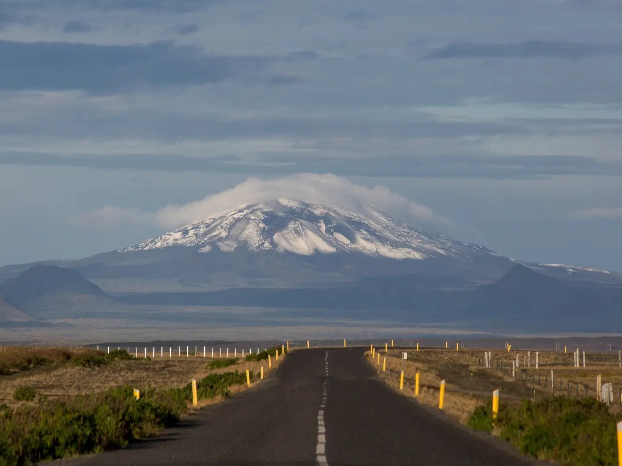 A long road leads to a majestic snow-capped mountain under a cloudy sky. The landscape is vast and barren, creating a serene, isolated atmosphere.