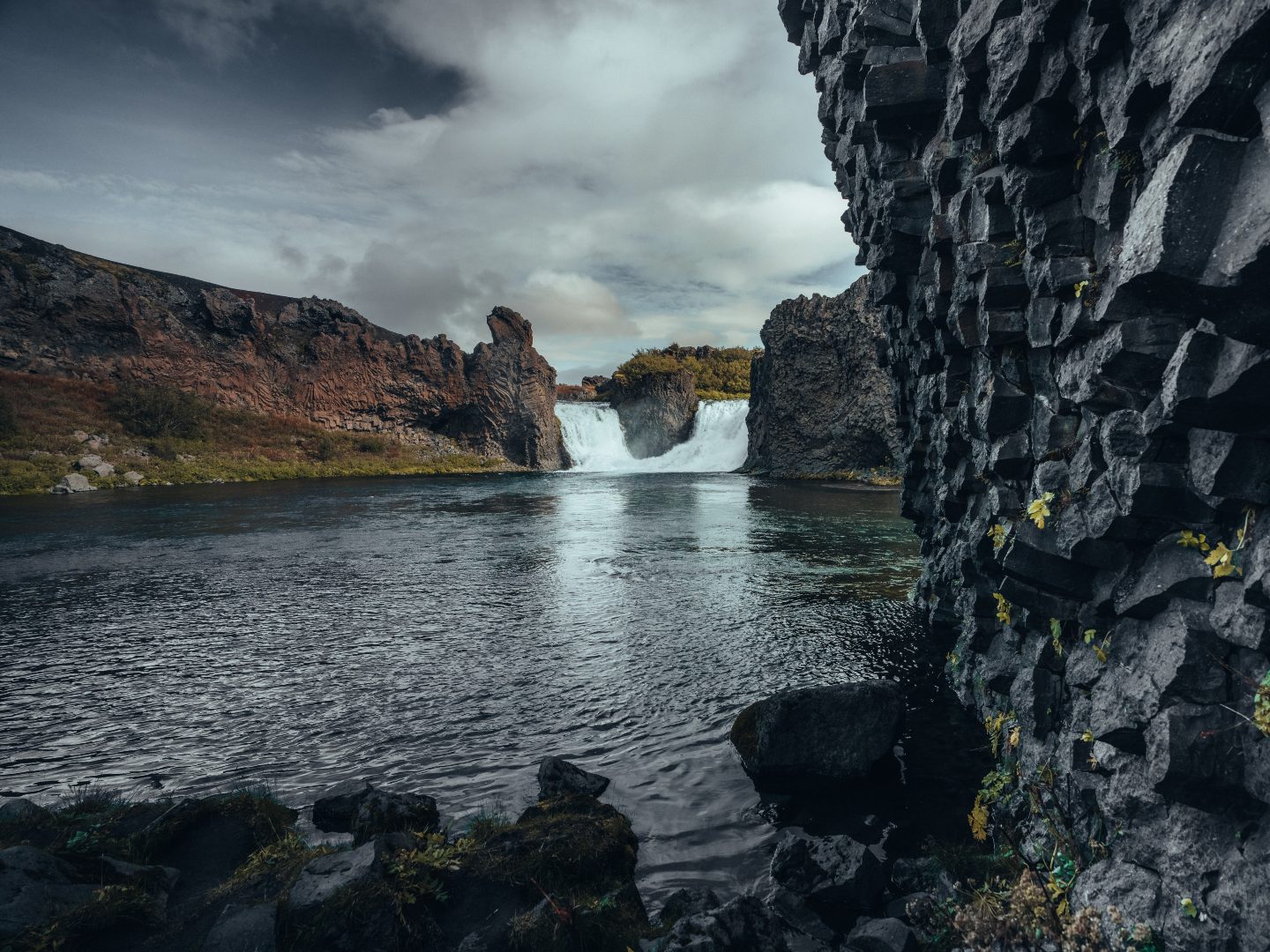 Rocky waterfall scene with jagged basalt cliffs on the right, cascading water in the center, and a calm river under cloudy skies, creating a serene atmosphere.