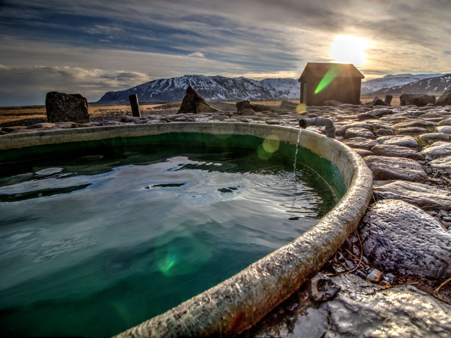 A tranquil hot spring with steaming water in a stone pool, surrounded by a rocky landscape. A small wooden hut and snowy mountains are visible under a setting sun.