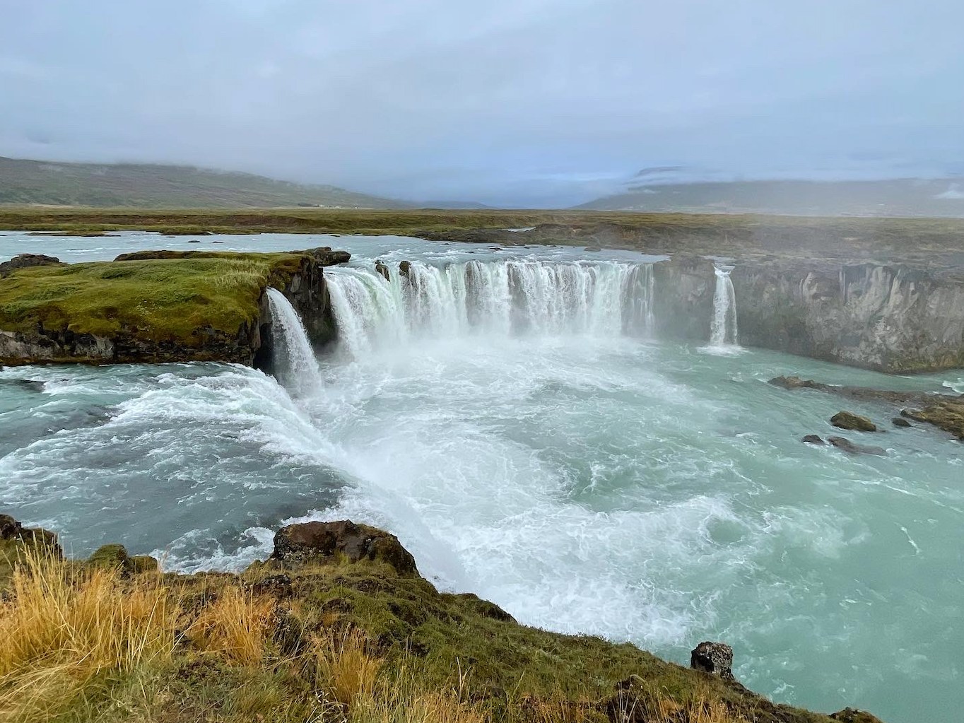 Powerful waterfall cascading over a rocky cliff into a turquoise river, surrounded by lush green grass under a misty, overcast sky. Tranquil yet dramatic.