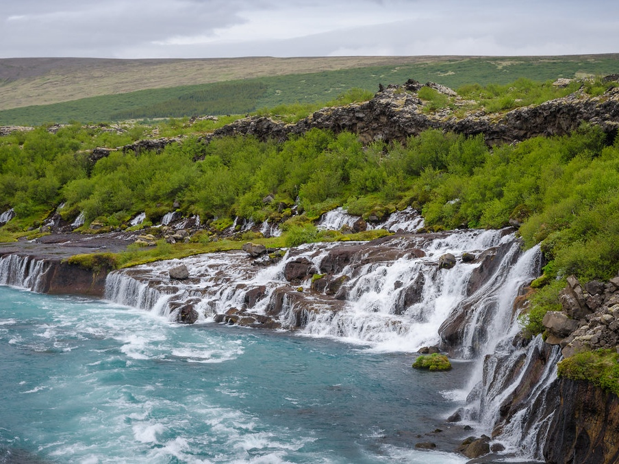 A lush green landscape with a cascading waterfall flows into vibrant blue waters. Rocky cliffs and greenery line the riverbank under a cloudy sky.