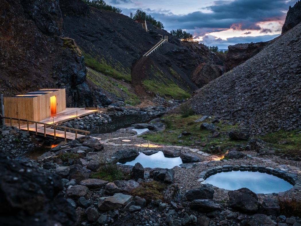 A serene landscape at dusk features a wooden cabin with warm lighting near rocky hot springs. A winding staircase leads up a dark, rugged hill.