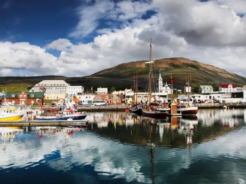 Scenic harbor view with colorful buildings and boats reflecting on calm water, under a dramatic cloudy sky with a large, grassy hill in the background.