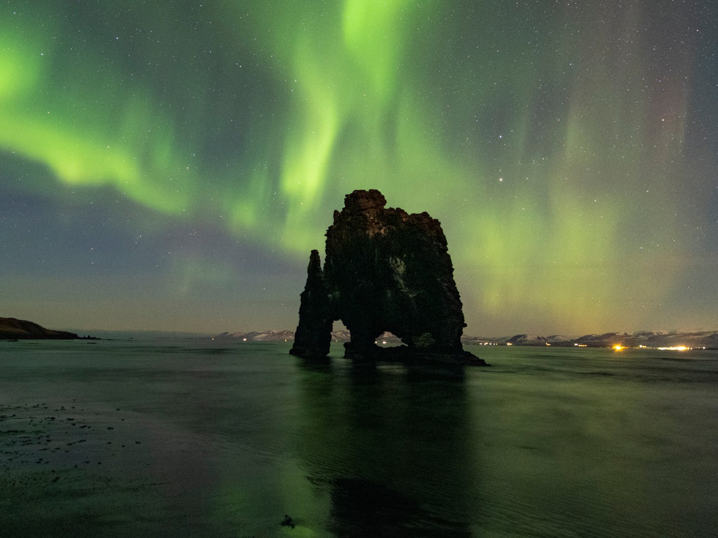 Northern Lights illuminate the night sky over a rocky sea stack formation. The vibrant green aurora reflects on calm water, creating a serene atmosphere.