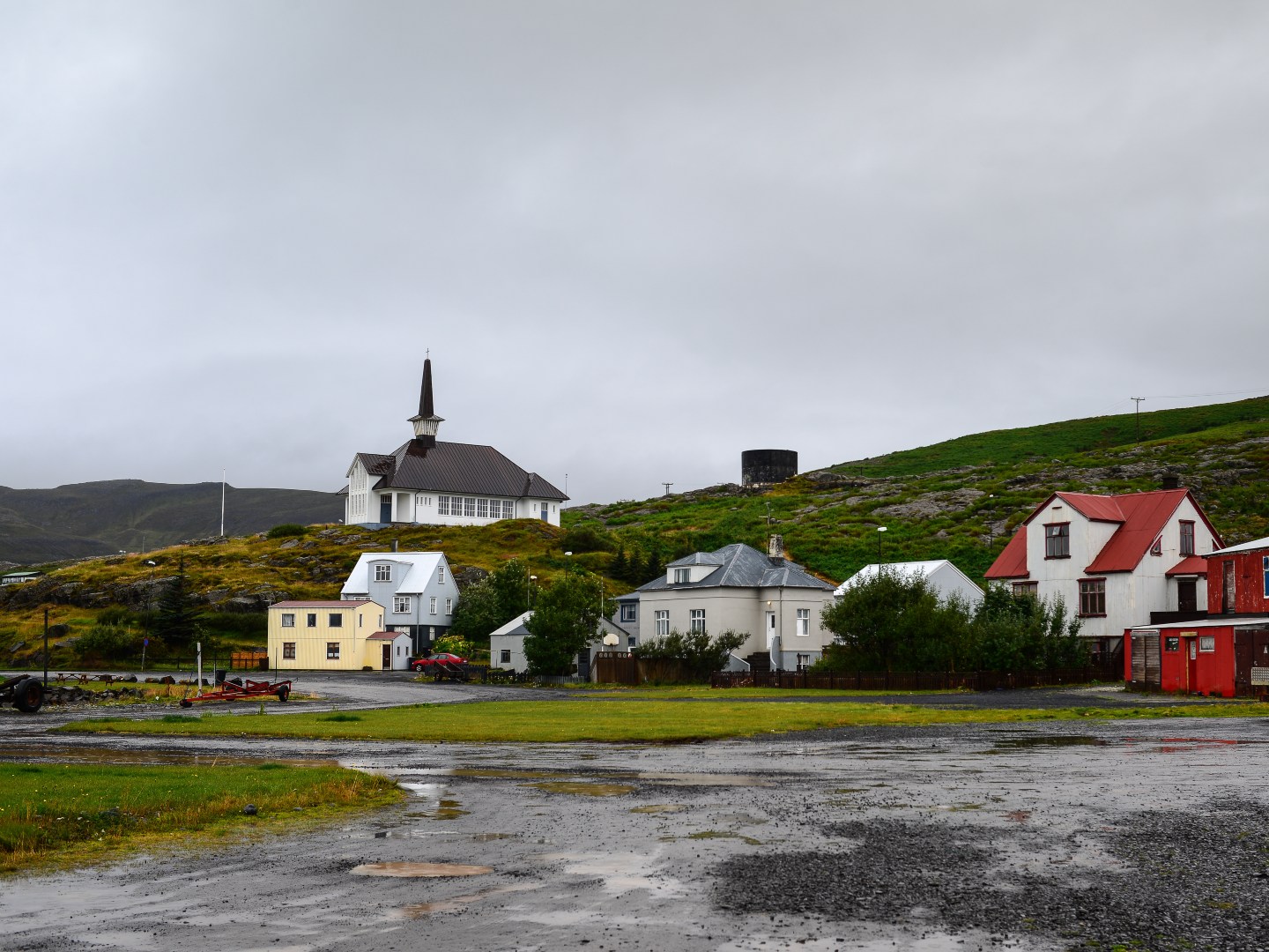 A small village under a cloudy sky, featuring a white church with a steeple on a hill, surrounded by colorful houses and lush green landscape.
