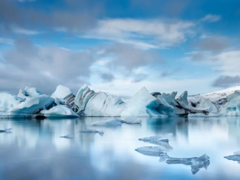Serene landscape of icebergs floating in calm, reflective water under a partly cloudy sky. The scene conveys a sense of tranquility and cold beauty.