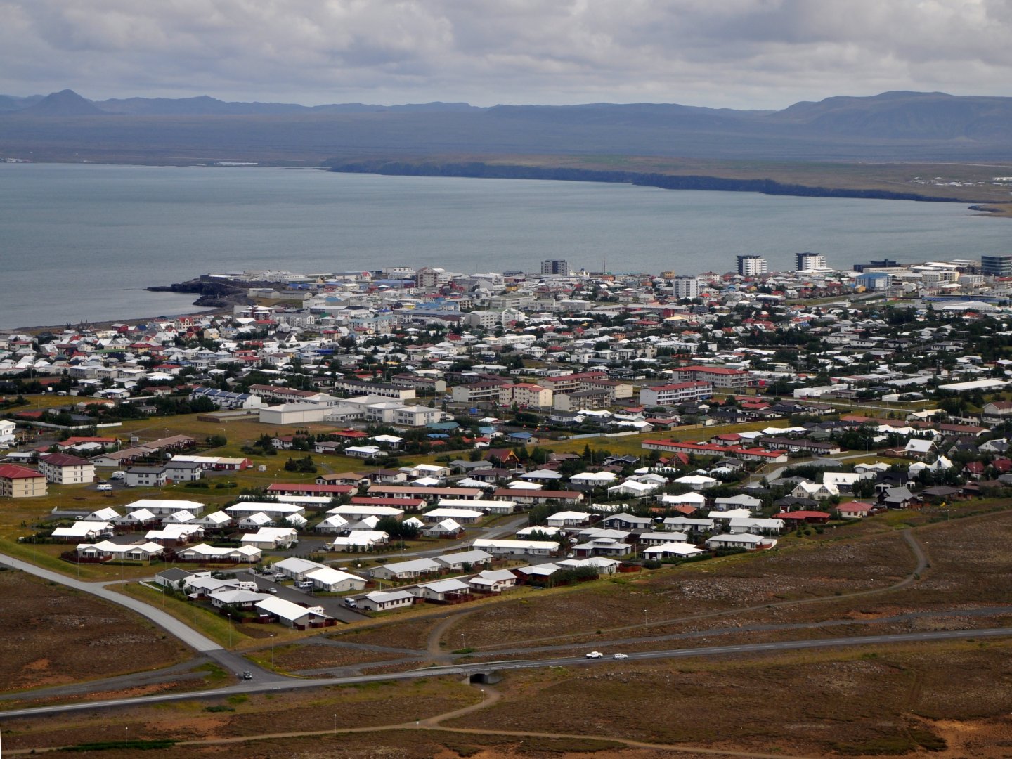 Aerial view of a coastal city with densely packed buildings and housing, overlooking a calm bay. Cloudy sky and distant hills create a serene atmosphere.