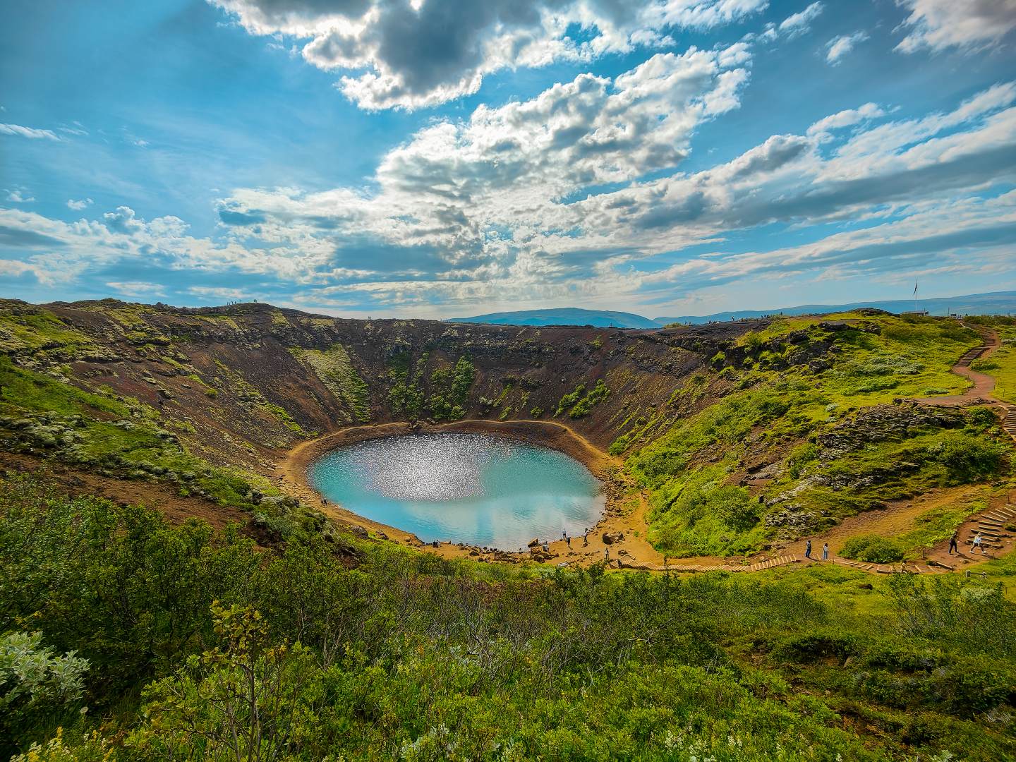 Scenic crater lake with shimmering blue water, surrounded by lush green vegetation and rocky terrain under a partly cloudy sky, conveying tranquility.