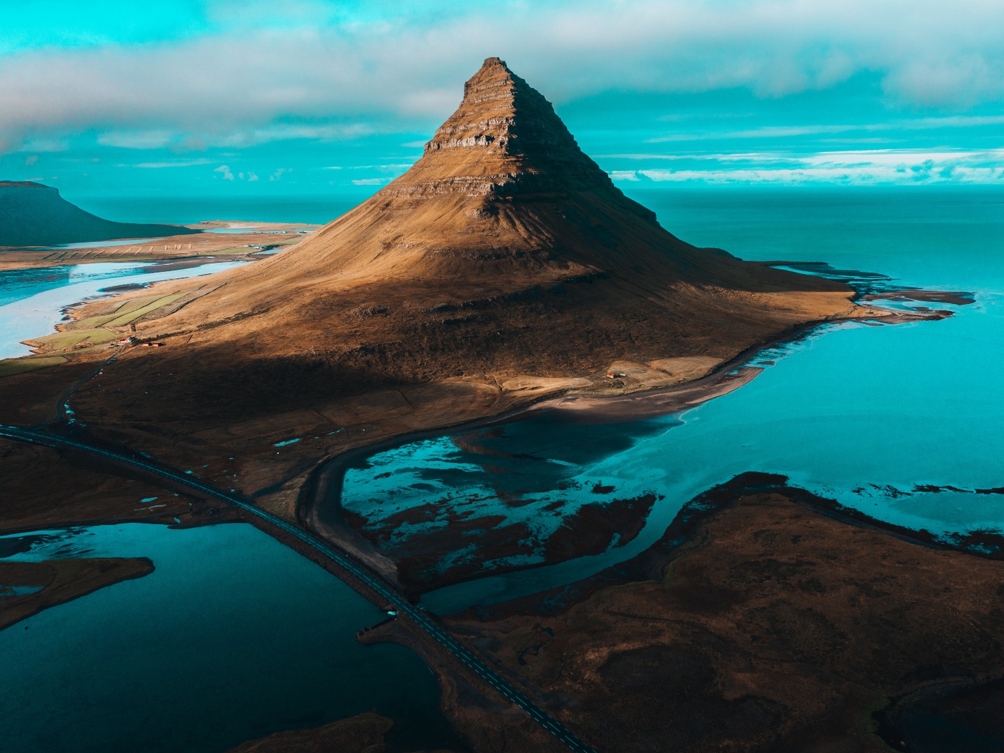 A conical mountain stands majestically surrounded by blue water and brown land under a vibrant blue sky, conveying a serene and dramatic landscape.