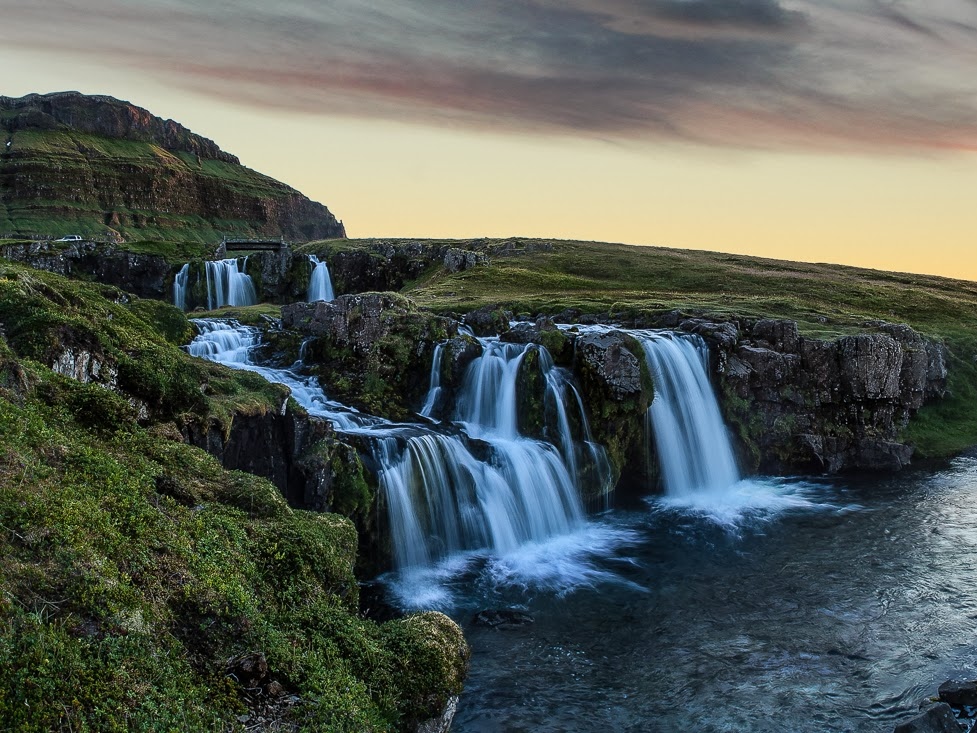 Scenic view of tranquil waterfalls cascading over rocky cliffs, surrounded by lush greenery. The sky above glows with a soft sunset, evoking calmness.