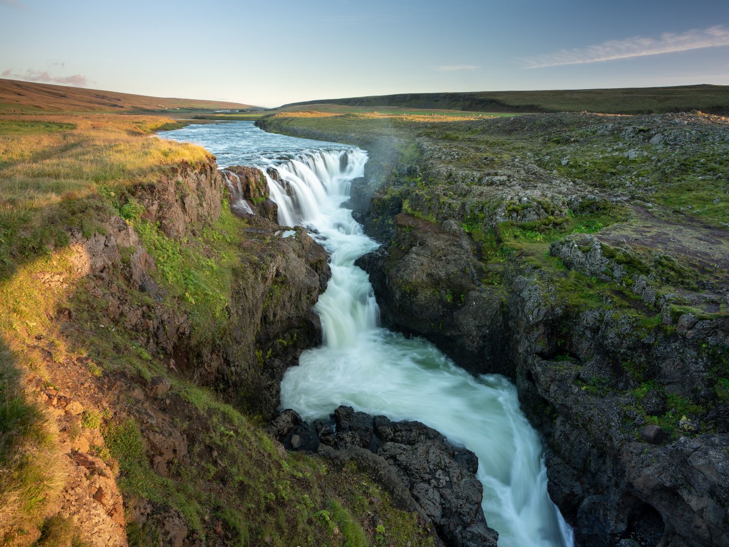 A serene waterfall cascades through a rocky gorge, surrounded by lush green grass under a bright blue sky, creating a peaceful natural scene.