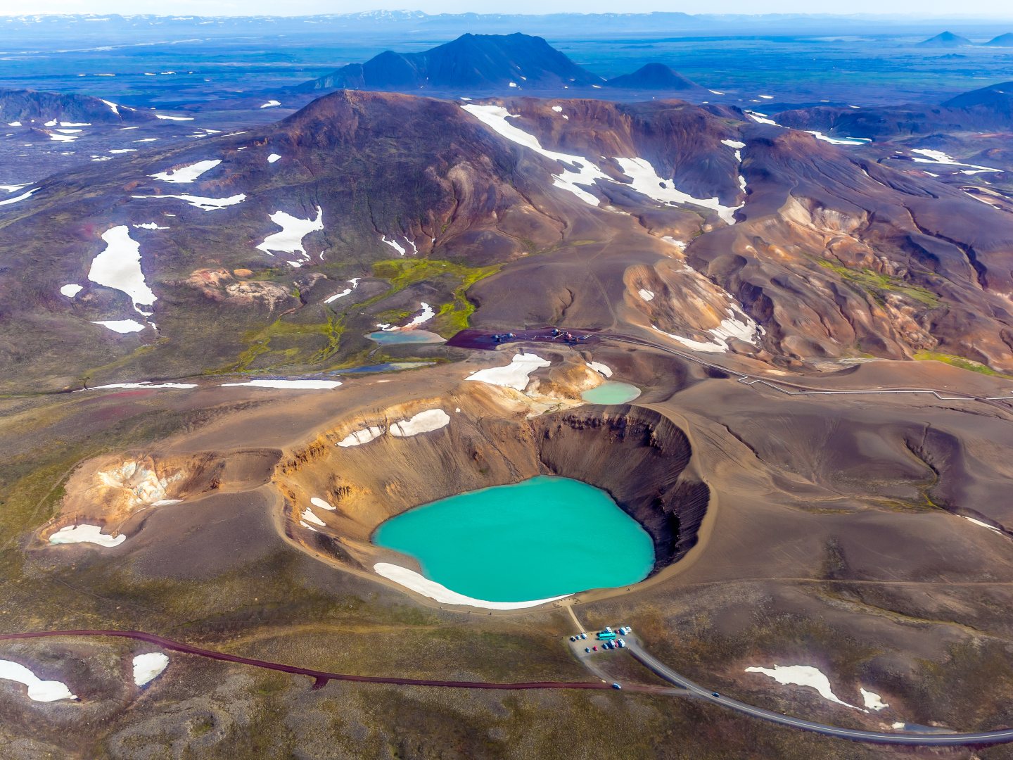 Aerial view of a vivid turquoise crater lake surrounded by rocky, snow-dotted mountains. Sparse vegetation adds contrast, creating a serene landscape.