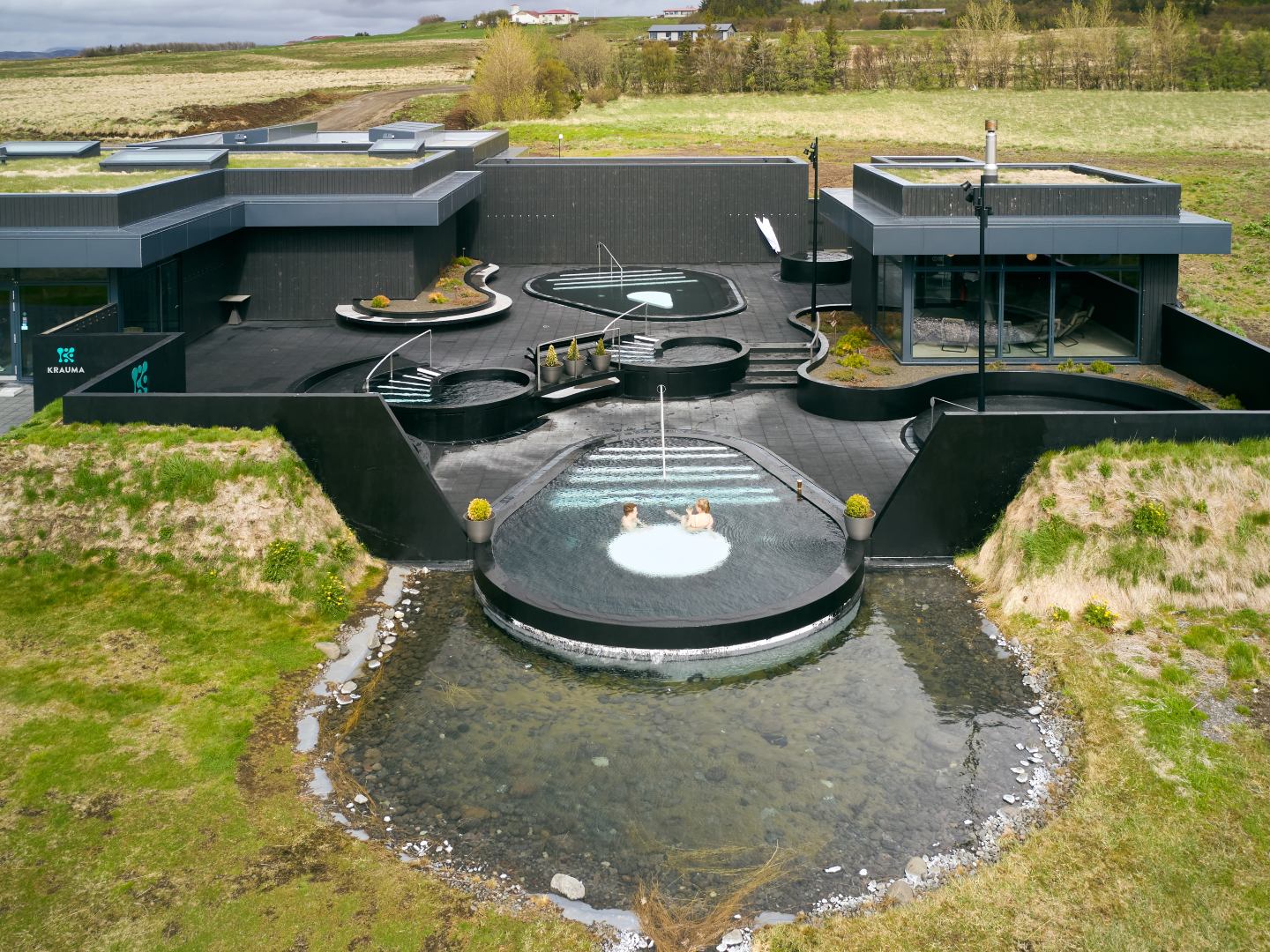 A modern spa with sleek black architecture sits in a rural landscape. Two people relax in a circular hot tub, conveying tranquility and leisure.