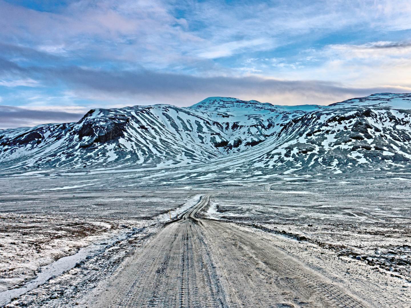 A snowy dirt road stretches toward snow-capped mountains under a partly cloudy blue sky. The scene conveys a sense of isolation and tranquility.