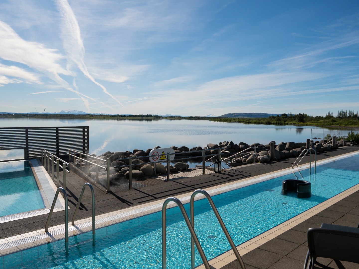 Outdoor pool with clear blue water and metal railings, overlooking a serene lake surrounded by rocks. The sky is clear blue with wispy clouds.