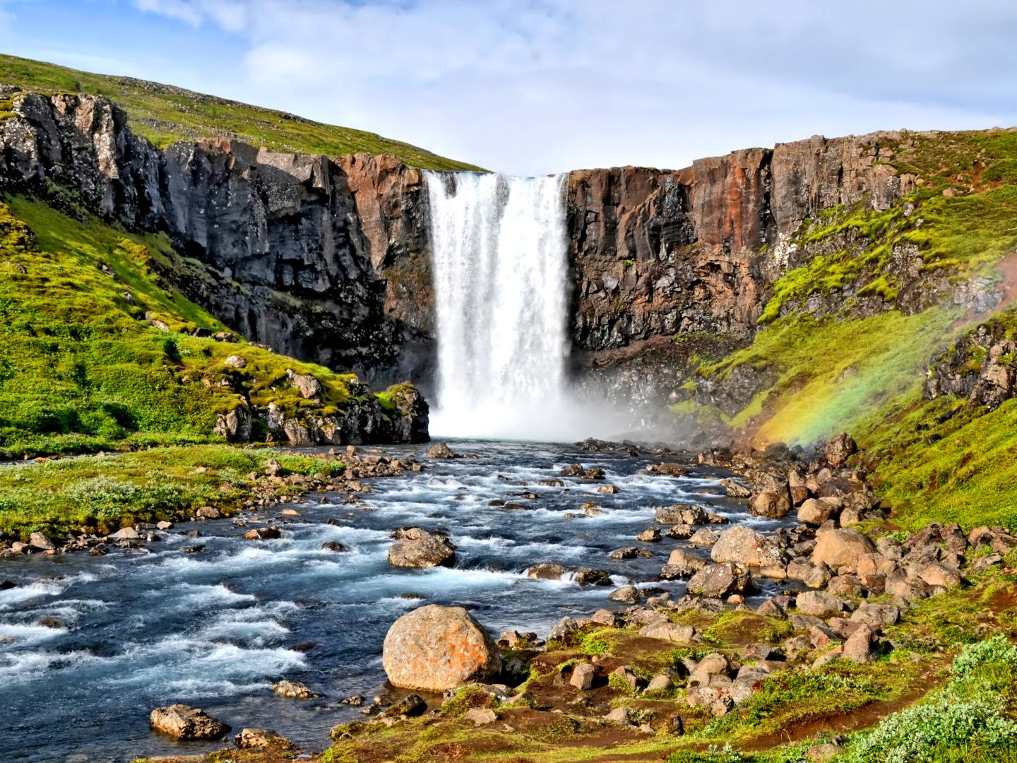 Majestic waterfall cascades over rocky cliffs into a flowing stream, surrounded by lush greenery. A faint rainbow arches over the right side, exuding serenity.