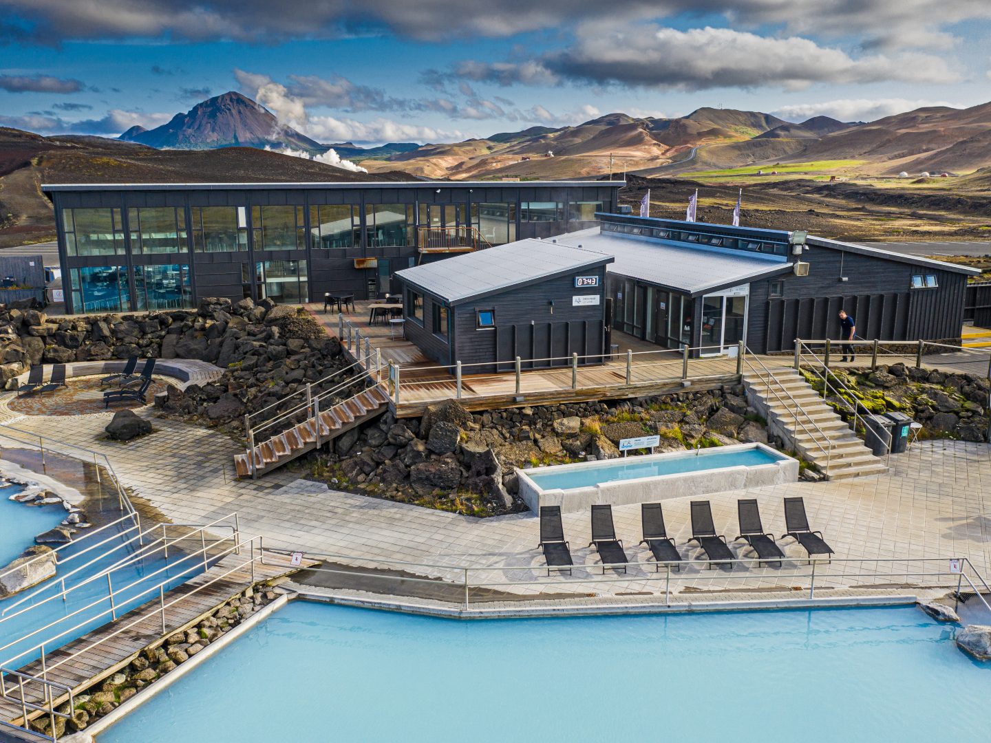 A modern geothermal spa with steaming blue water pools, surrounded by black lava rocks. In the background are glass buildings and scenic mountains.