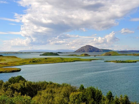 Serene landscape of a lake with scattered grassy islets under a partly cloudy sky. A distant mountain enhances the tranquil scene.
