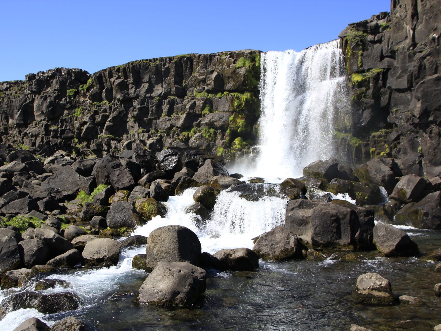 A serene waterfall cascades over dark, rocky cliffs into a clear stream. Green moss accents the rocks, and the scene is set against a bright blue sky.