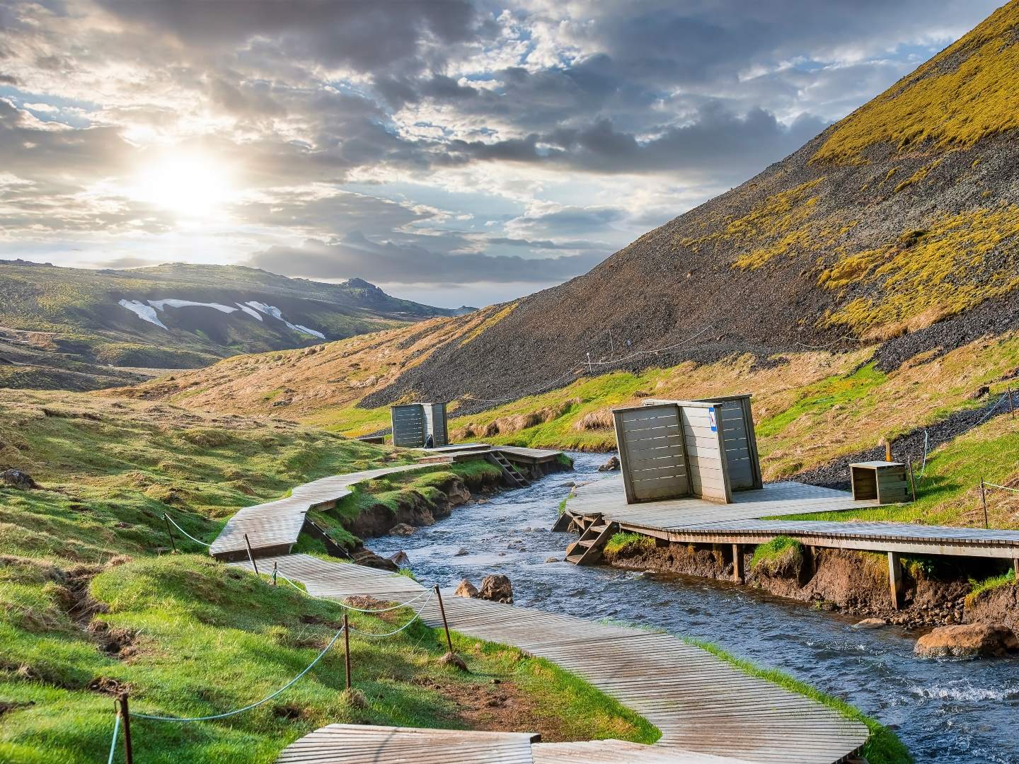A winding wooden pathway follows a serene stream through a green valley at sunrise. Cloudy skies and surrounding hills create a tranquil, natural atmosphere.