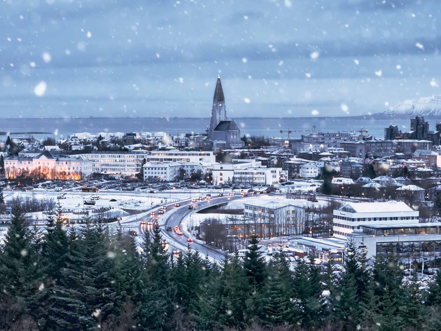 Snow falls softly over a wintery cityscape with a prominent church tower. Warm lights glow from buildings, and trees frame the serene scene.
