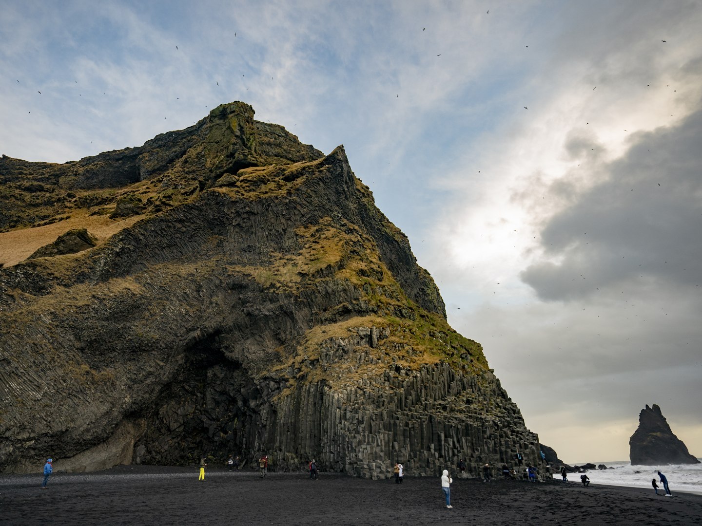 Dramatic coastal cliff with hexagonal basalt columns at Reynisfjara Beach, Iceland. Dark sand, stormy sky, scattered visitors, rugged landscape.