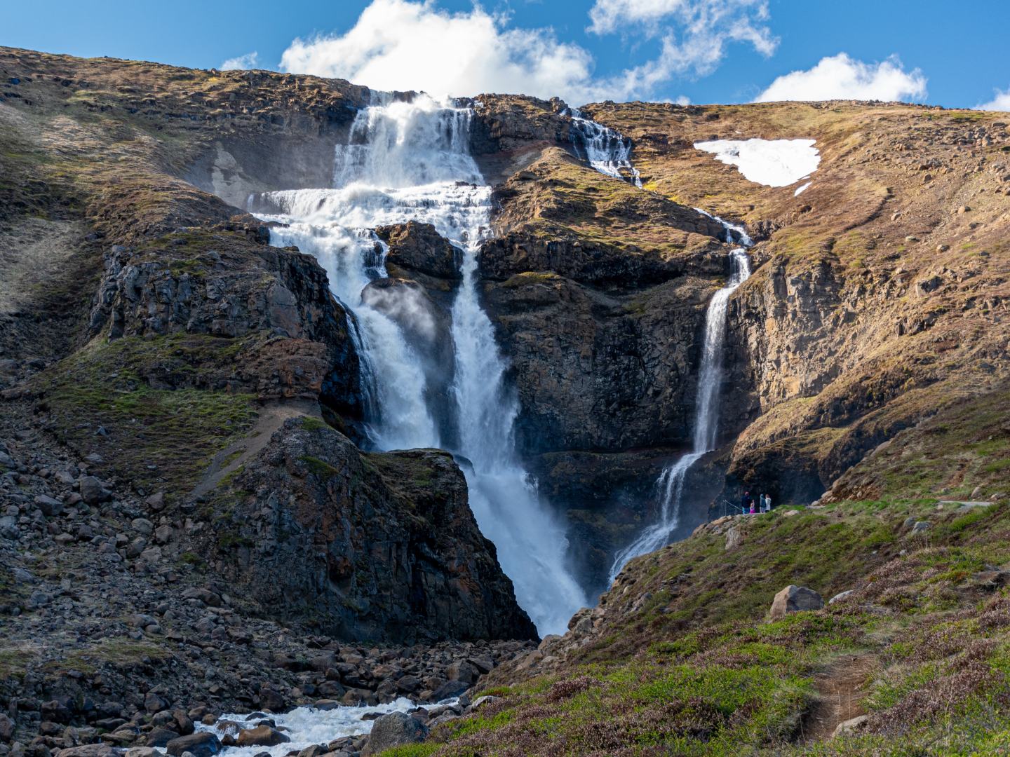 Majestic waterfall cascading down rugged cliffs, surrounded by rocky terrain and sparse greenery under a clear blue sky with a few clouds.