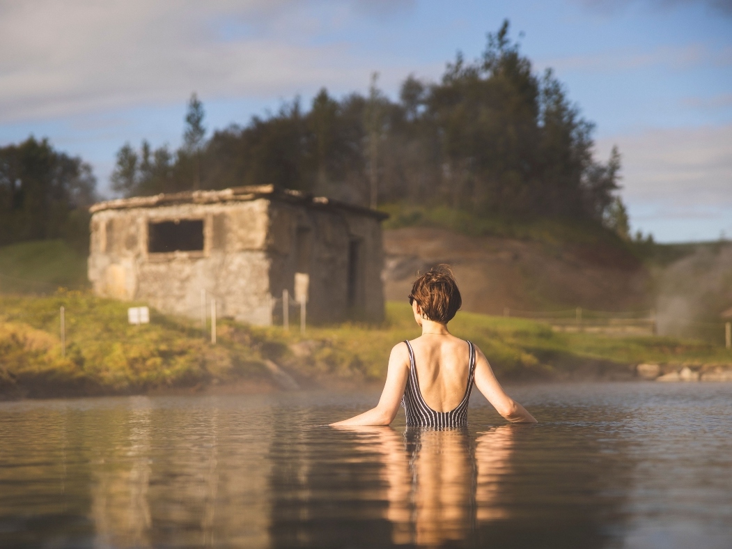 A woman in a swimsuit wades in a hot spring with an old stone building in the background, surrounded by mist and trees, evoking tranquility.