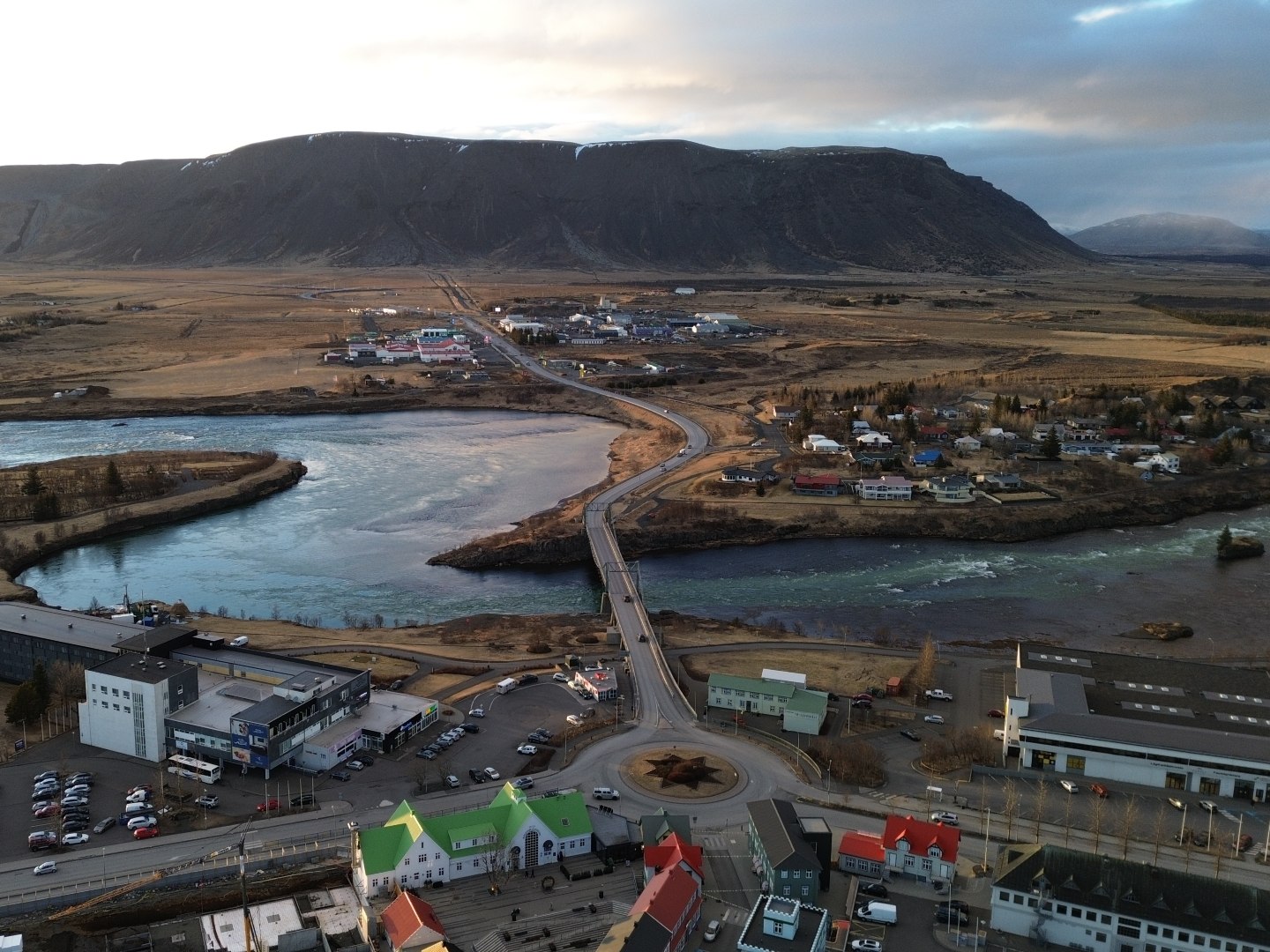 Aerial view of a small town with a winding river and bridge. Colorful houses in the foreground, with barren hills in the distance under a cloudy sky.
