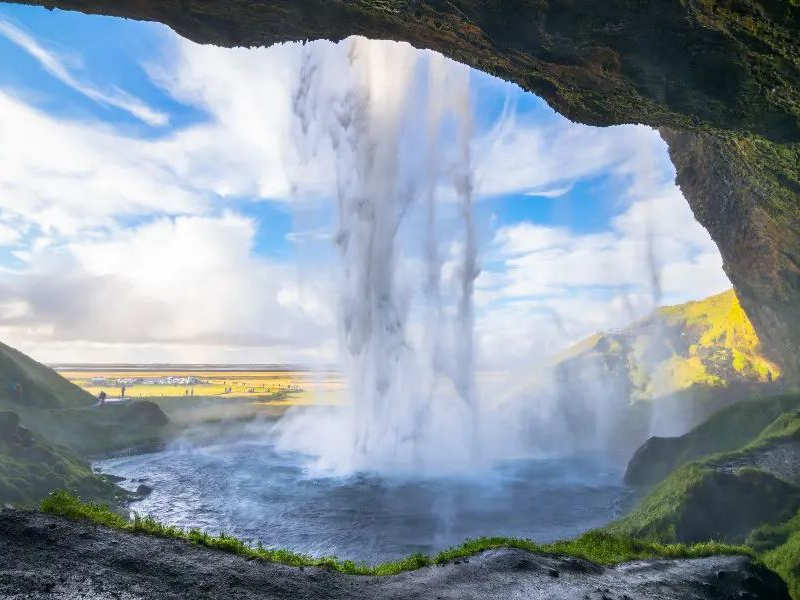View from behind a waterfall with clear blue skies. Water cascades down into a pool, surrounded by lush greenery, creating a serene and majestic scene.