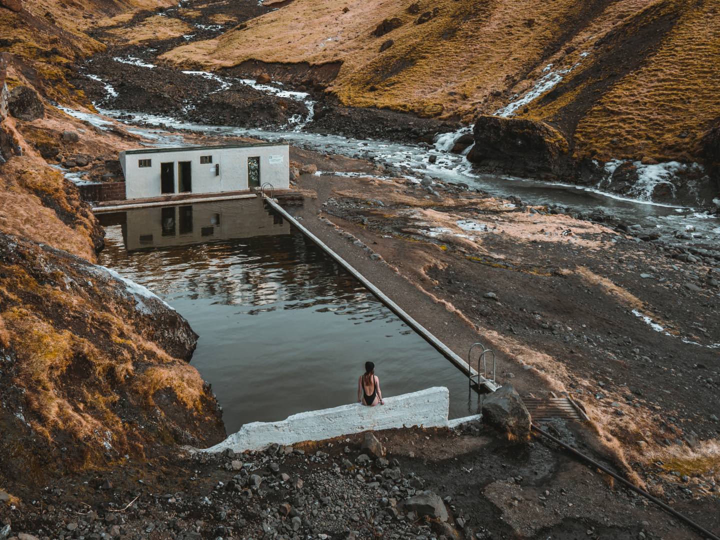 A person sits by a remote outdoor pool amid rugged hills and a flowing stream, with a small building nearby. The scene feels tranquil and isolated.