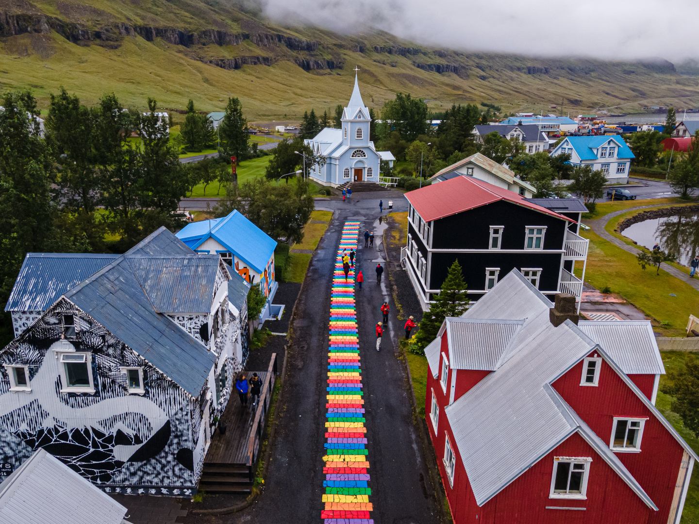 Aerial view of a picturesque village with colorful houses lining a street painted in rainbow stripes. A quaint, white church sits at the end, surrounded by lush greenery and misty hills. The scene is vibrant and inviting.