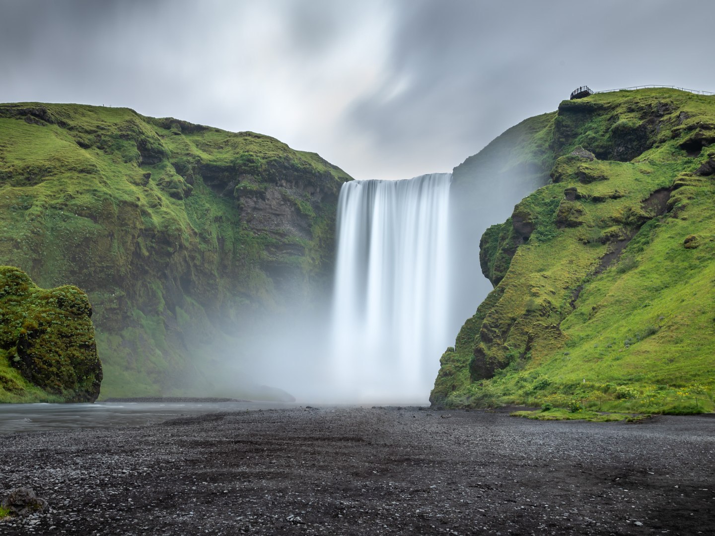 A majestic waterfall cascades down a wide cliff between lush green hills under a cloudy sky, with mist rising from the water and a serene atmosphere.