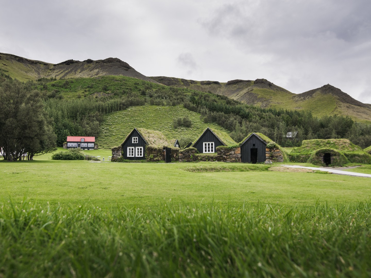 Green landscape with traditional black turf houses featuring grass roofs, set against rolling hills and cloudy sky. A red house is visible in the distance.