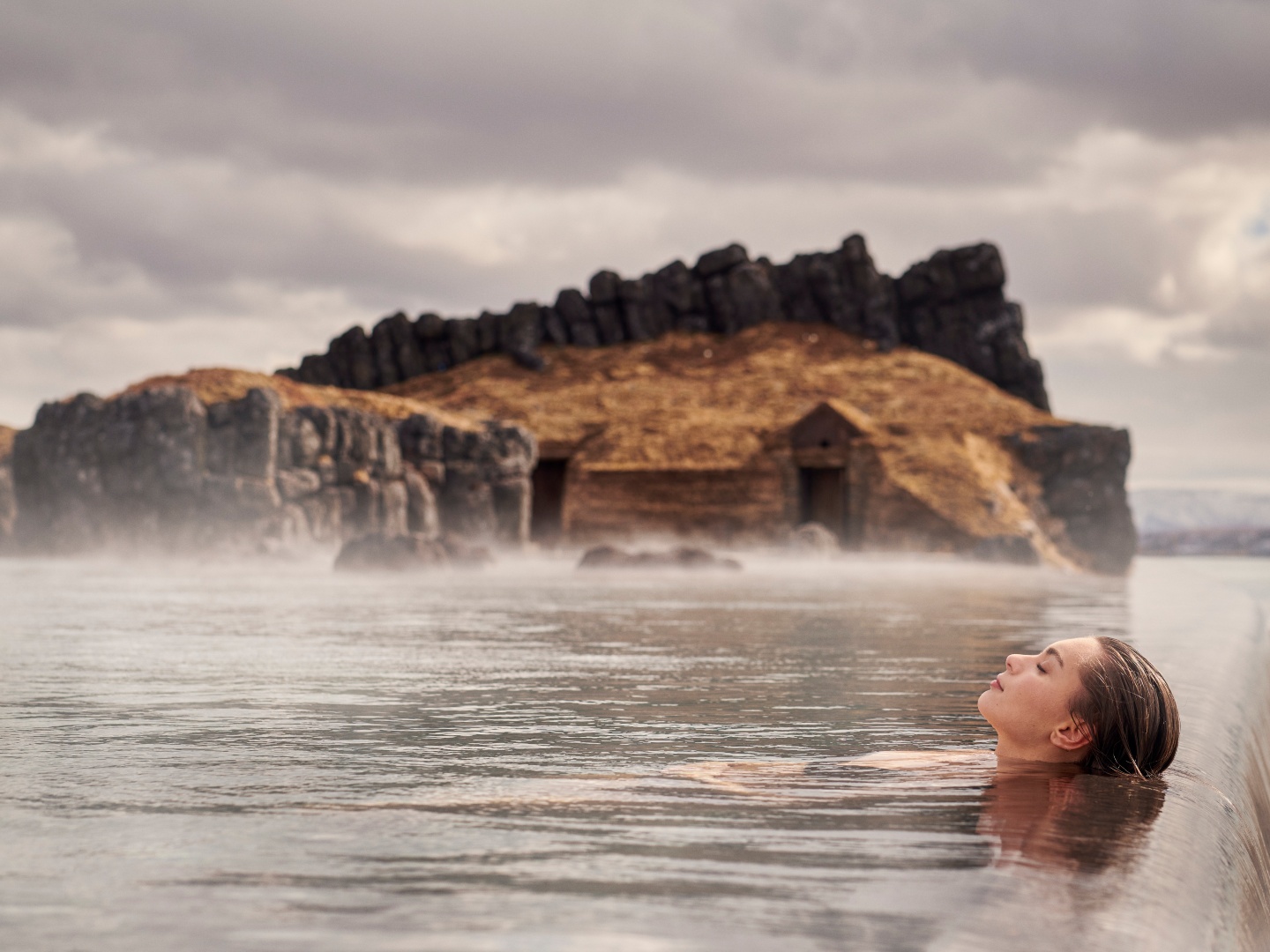A person relaxes in a serene geothermal pool under a cloudy sky, with rugged rocks and a mossy cabin in the background, evoking tranquility.