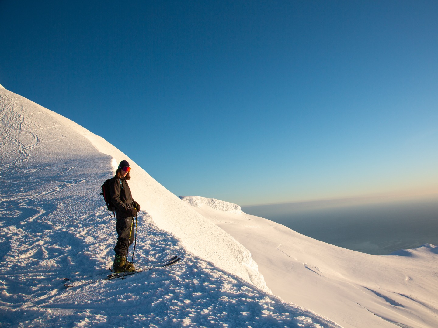 A skier in winter clothing stands on a snowy mountain slope under a clear blue sky. He gazes outward, conveying a sense of tranquility and adventure.