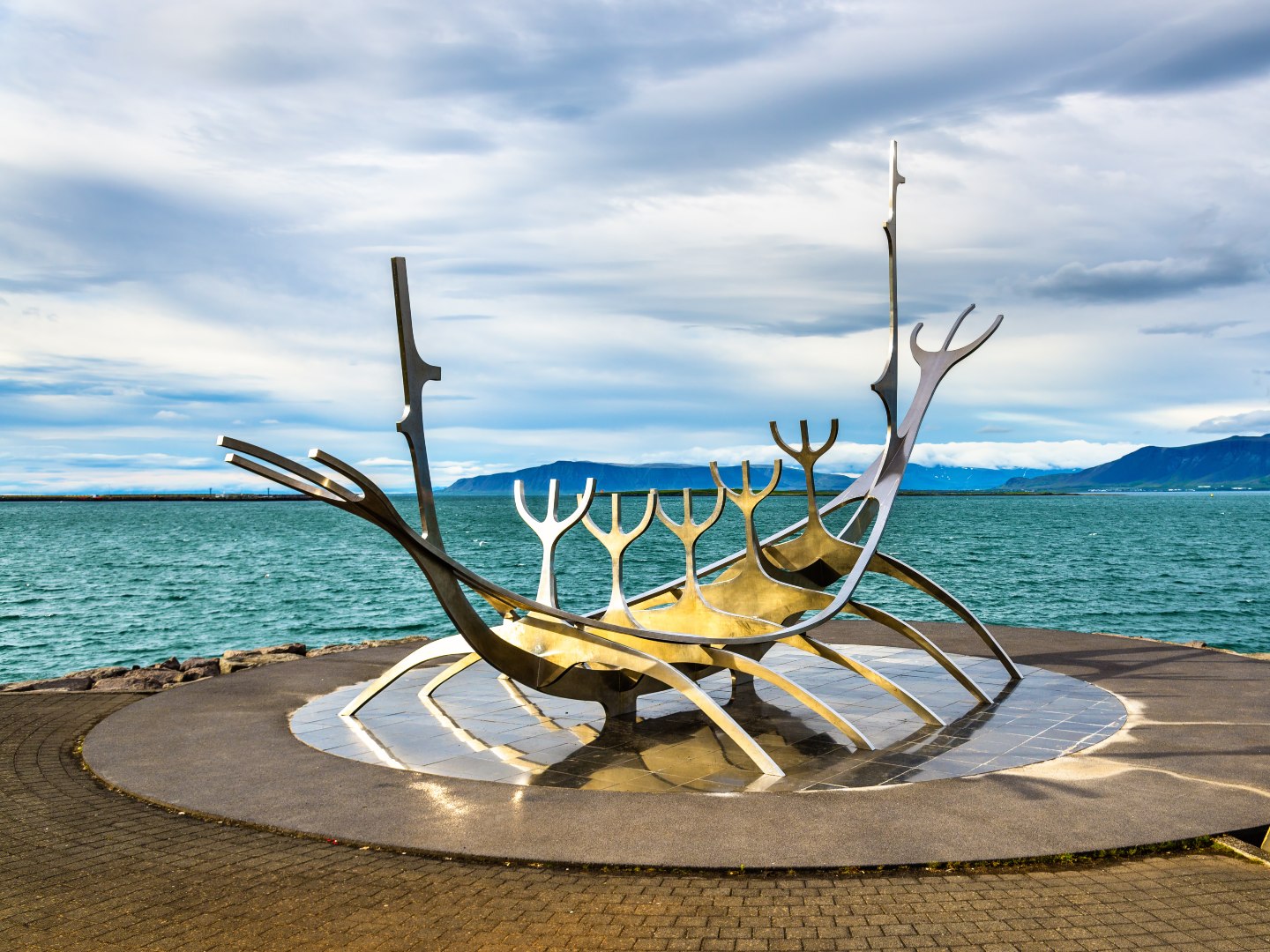 Sleek silver sculpture resembling a Viking ship, known as "Sun Voyager," by the sea with a cloudy sky and distant mountains. Calm and reflective atmosphere.
