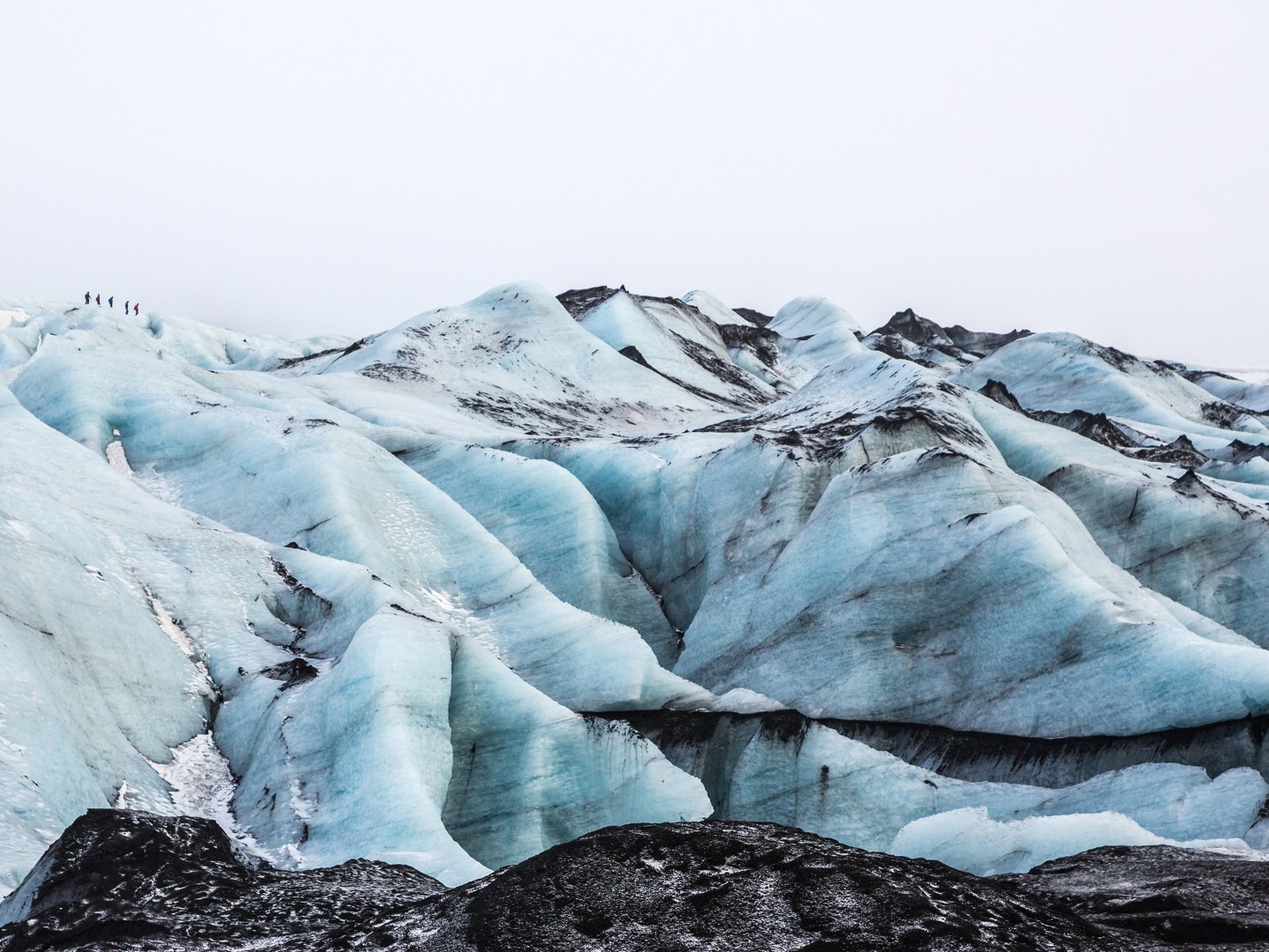 Snowy glacier landscape with swirling blue ice formations and dark streaks of volcanic ash. Small figures near the top suggest scale and grandeur.