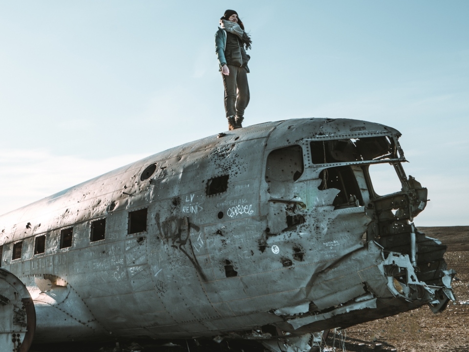 A person stands confidently atop a weathered, graffiti-covered airplane wreck in a barren landscape under a clear blue sky, conveying a sense of adventure.