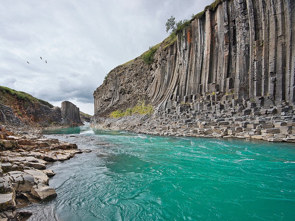 A turquoise river flows through a canyon flanked by towering, columnar basalt cliffs. The sky is cloudy, and birds can be seen flying overhead.