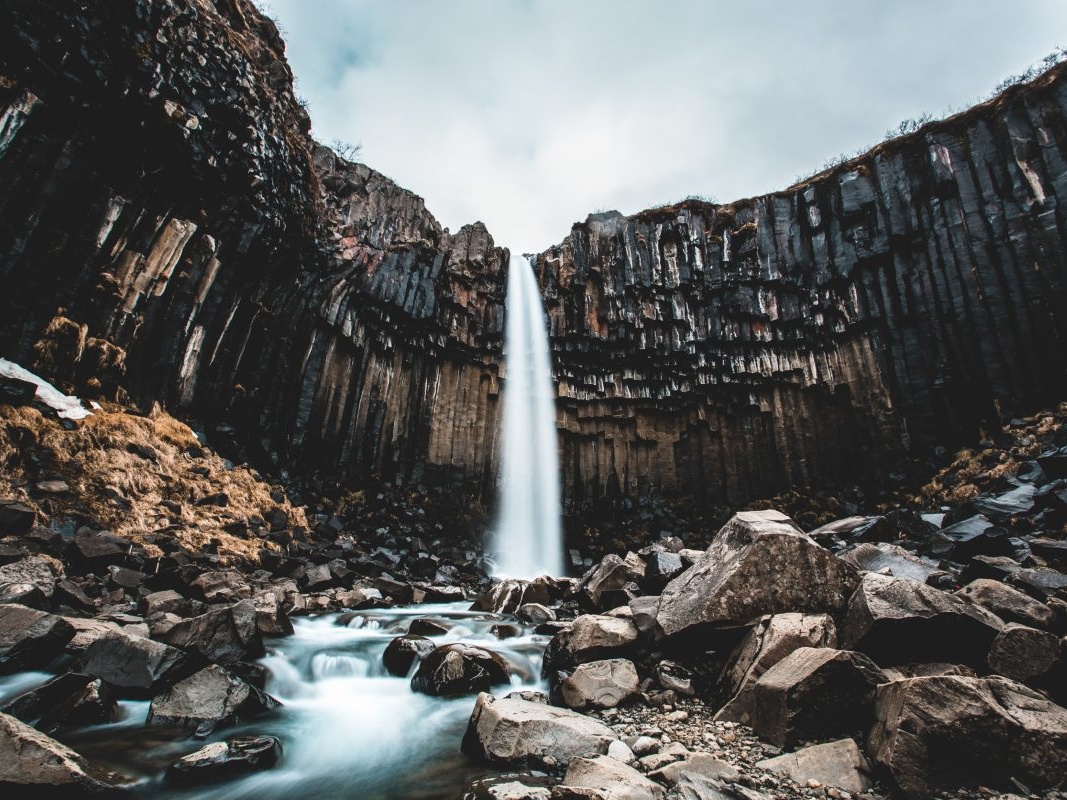 Tall waterfall cascading over dark basalt columns, surrounded by rugged rocks and a cloudy sky. The scene conveys a dramatic, natural beauty.