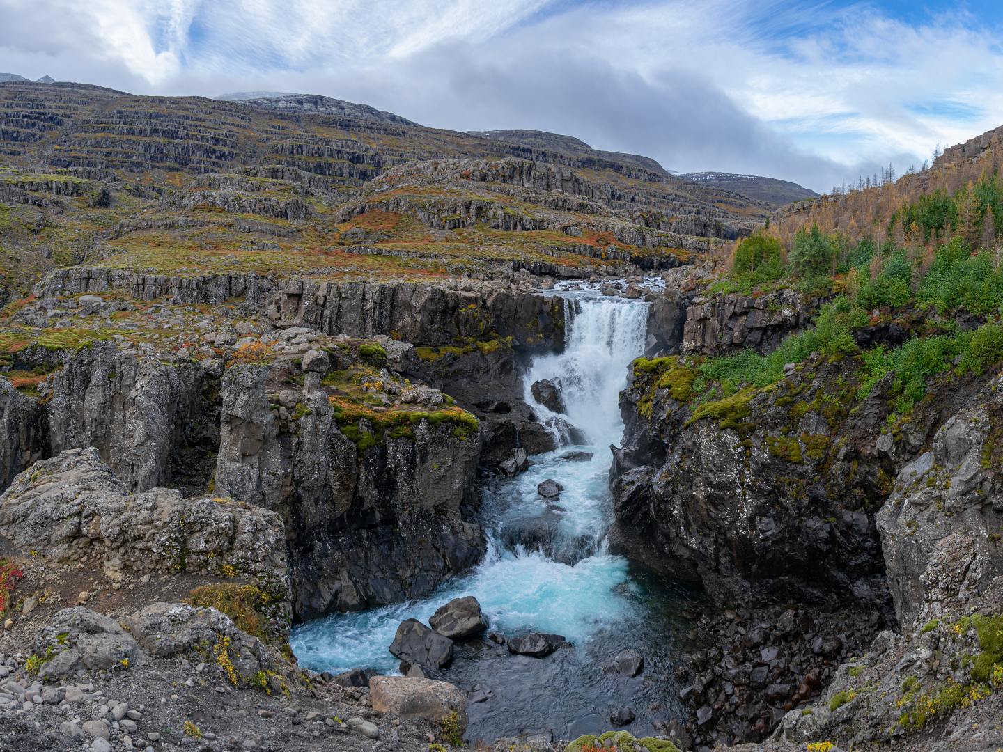 A serene waterfall cascades through rugged cliffs with lush greenery. The sky is partly cloudy, creating a tranquil landscape scene.