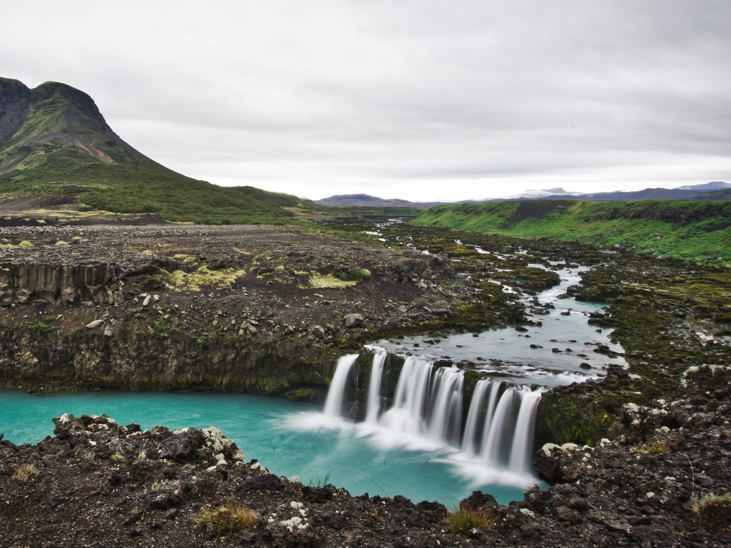 A serene landscape of a small waterfall cascading into a turquoise river, surrounded by rocky terrain and green hills under a cloudy sky. Peaceful ambiance.