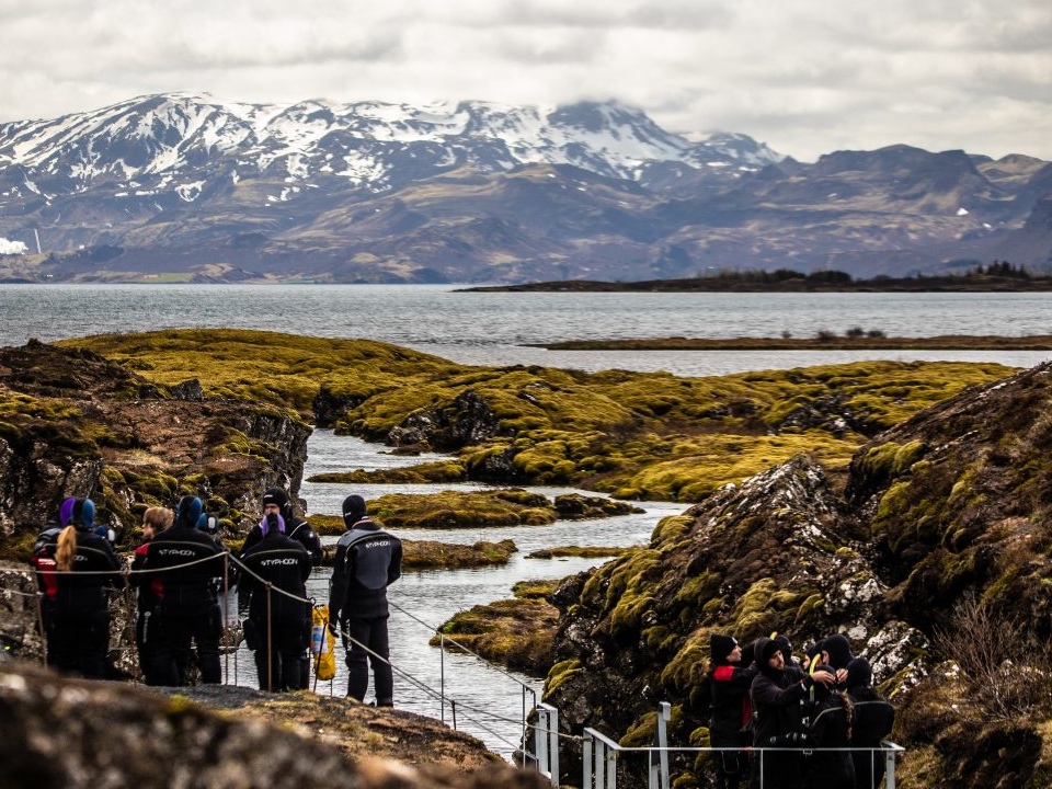 People in wetsuits stand by a narrow, moss-covered fissure leading to a lake, with snow-capped mountains in the background under a cloudy sky.
